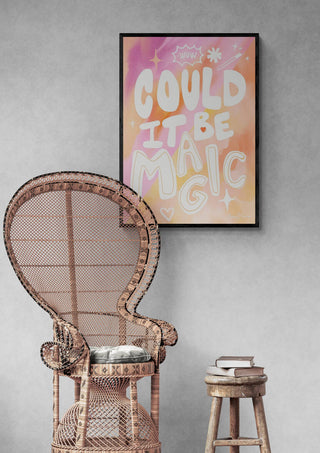 Framed "Could it Be Magic" Peach Pink poster on a gray wall above a wicker chair and small stool with books.