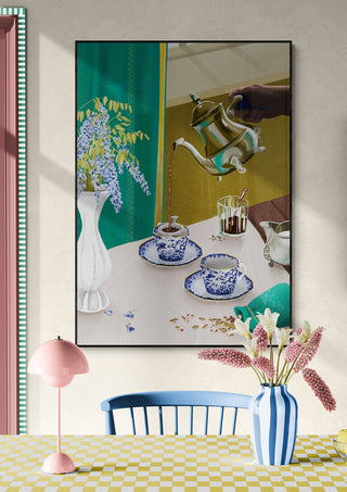 Framed art print of a blue-patterned tea set, flowers, and hand pouring tea; displayed in a modern dining room.