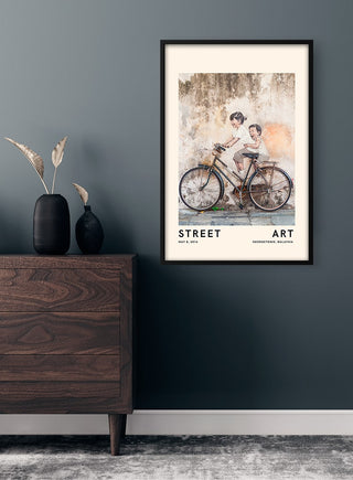 Framed poster of Malaysian street art featuring two children on a bicycle, hung above a wooden dresser.