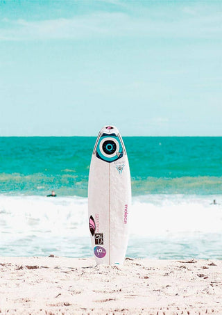 White surfboard standing upright on sandy beach with blue ocean waves in the background.