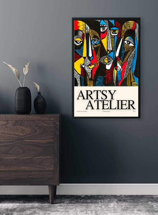 Framed This is Artsy Atelier Plakat with colorful abstract faces, hung on a dark wall above a wooden cabinet.