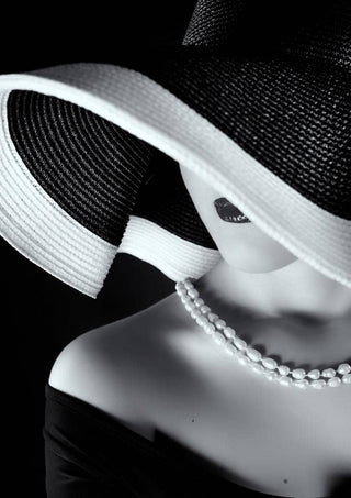 Black and white portrait of a woman in a wide-brim hat, pearl necklace, and off-shoulder dress.