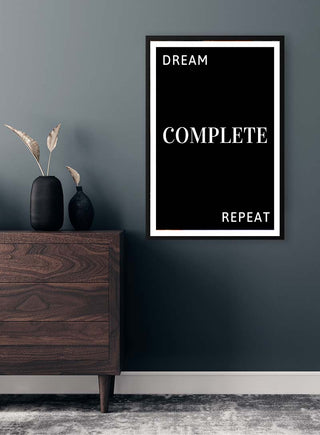 Framed Dream Complete Black poster on a dark wall above a wooden dresser with decorative vase and dried plants.