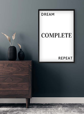 Minimalist "Dream Complete Repeat" print in black frame on dark wall, styled with modern wooden dresser and vase.