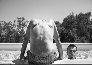 Black and white photo of a man in patterned shorts sitting poolside, another man's face emerging from the water.