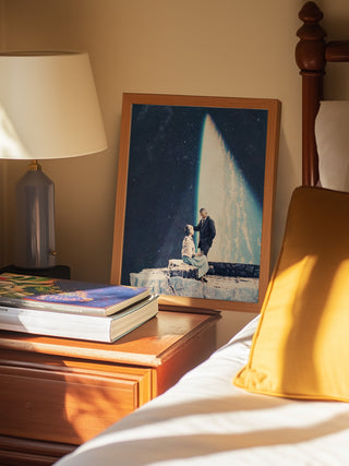 Framed "This Love" by Frank Moth art print on a bedside table next to lamp, books, and bed with yellow pillow.