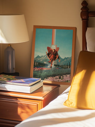 Framed Enemy by Frank Moth artwork on bedside table next to lamp, books, and pillows in a cozy bedroom setting.