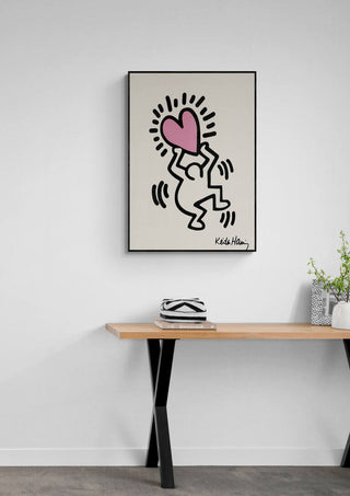 Framed Hearthead artwork by Keith Haring featuring a figure holding a pink heart, displayed above a wooden table.