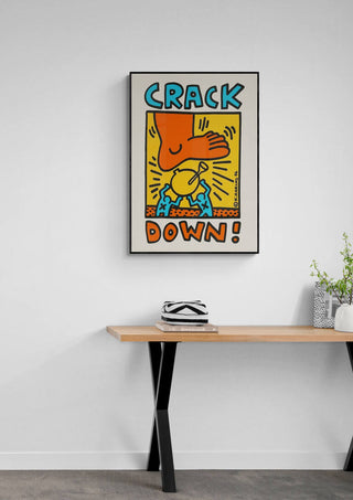 Framed "Crack Down" by Keith Haring hanging above a modern wooden desk with plants and books.