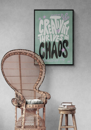 Framed "Creative Chaos - Baroo Bloom" art print above wicker chair and stool with books against gray wall.