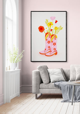 Framed art of pink cowgirl boots filled with colorful flowers, displayed above a modern sofa.
