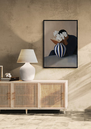 Framed "Flower Reading" by David Wolf displayed above a sideboard with a white lamp in a modern interior.