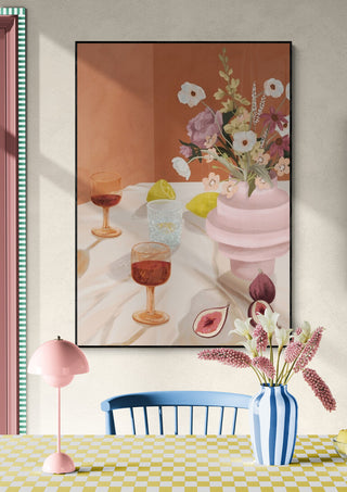 Framed art print of a summer table with drinks and flowers, above a pastel dining set in bright sunlight.