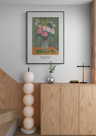Framed "Flowers In a Vase" by Henri Rousseau displayed above a modern wooden cabinet in a minimalist room.