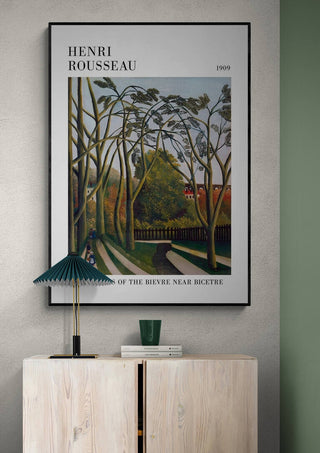 Framed print of "The Banks Of The Bier" by Henri Rousseau hanging above a light wood cabinet.