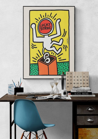 Lucky Strike Keith Haring art poster above a modern desk with books, calendar, and blue chair.