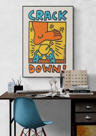 Framed "Crack Down" by Keith Haring displayed above a modern desk with books and decor.